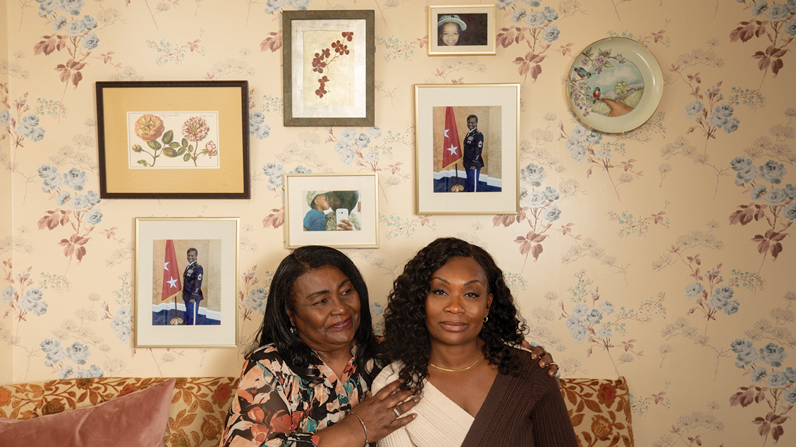 A Veteran and her mother sitting on a couch in front of floral wallpaper and a collage of portraits including some from her service.