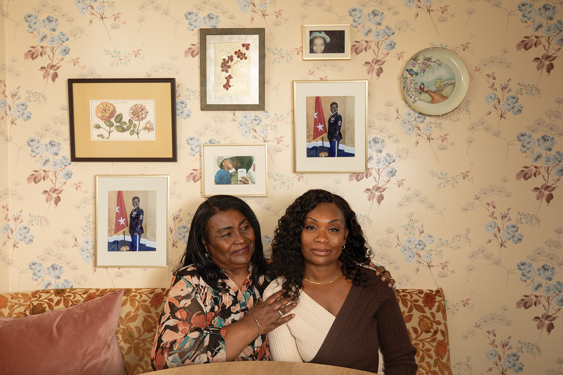 A Veteran and her mother sitting on a couch in front of floral wallpaper and a collage of portraits including some from her service.