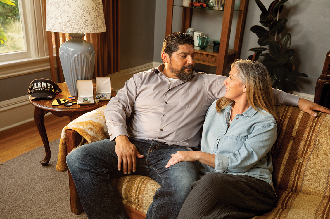 Veteran James and April sitting on the couch cozied up looking each other in the eye with an Army Veteran hat on the end table.