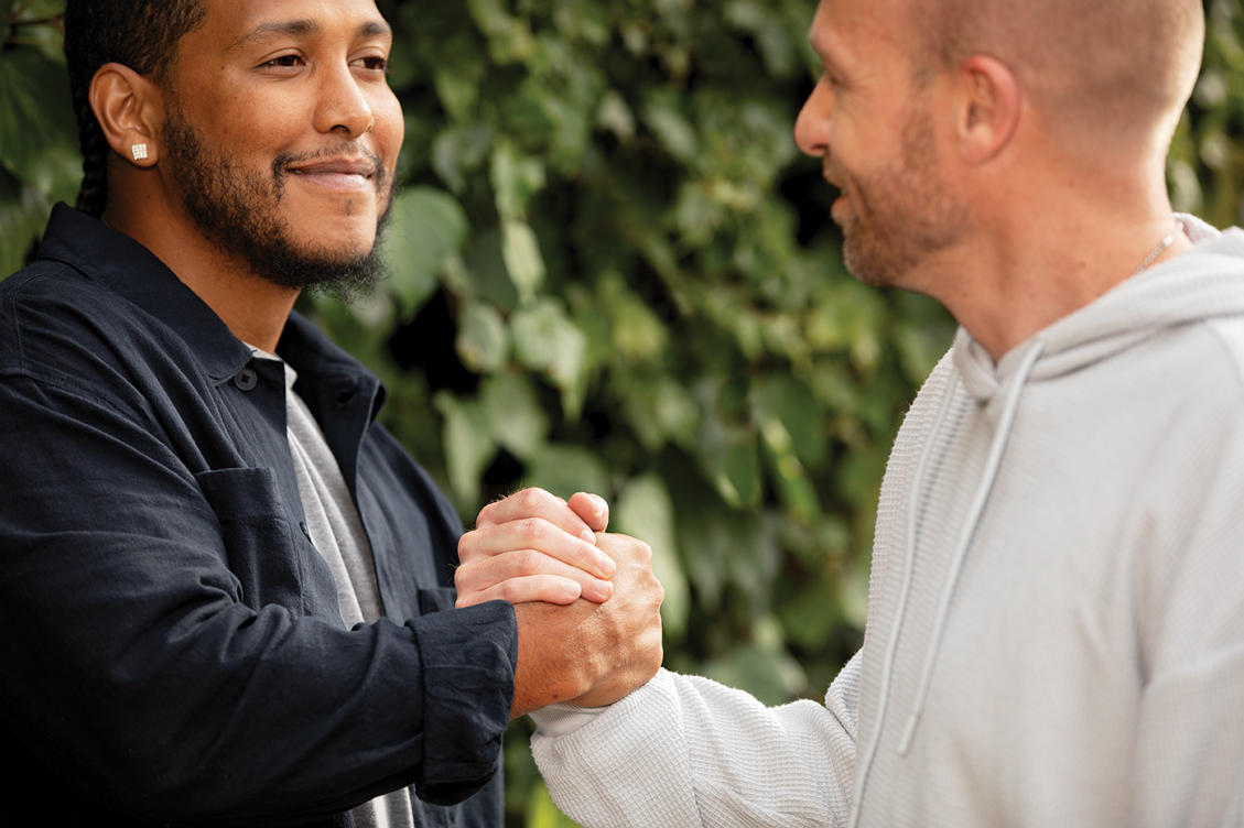 Two gentlemen, Sean and Josuha clasping hands and looking each other in the eye smiling outside.