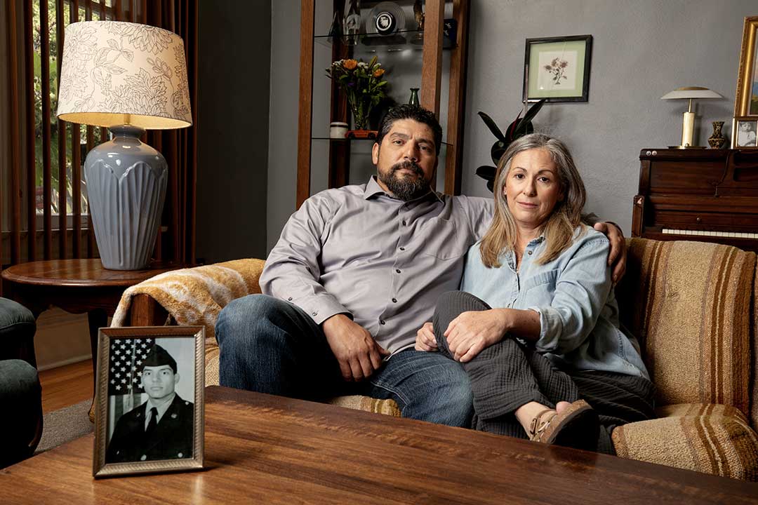 Veterans James and April sitting on the couch looking toward the camera with a photo of James in his Army uniform. 