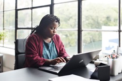 woman sitting at a desk by a window typing on a laptop