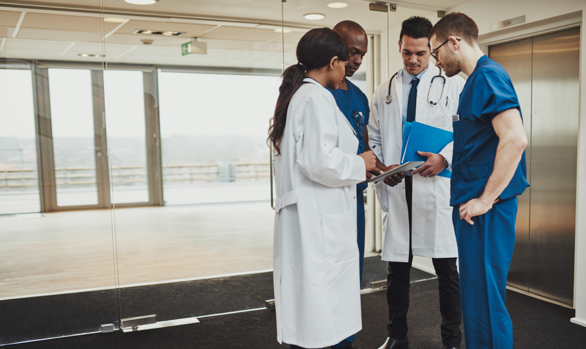 A group of four medical professionals, three men and one woman, consulting charts near an elevator bank.