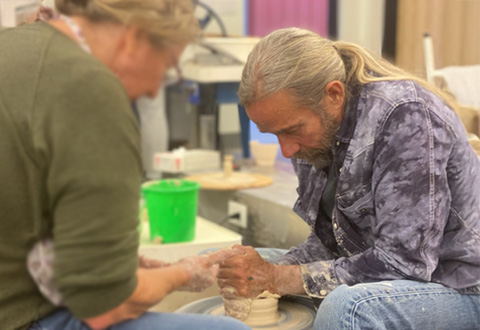 Man helps student shape clay on a potter’s wheel.