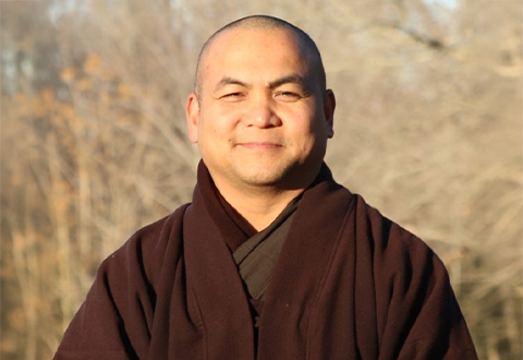 Man standing in front of trees in brown robe smiles for a picture