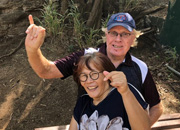 smiling man and woman sitting on bench in front of trees making hang loose hand gesture