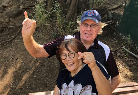 smiling man and woman sitting on bench in front of trees making hang loose hand gesture