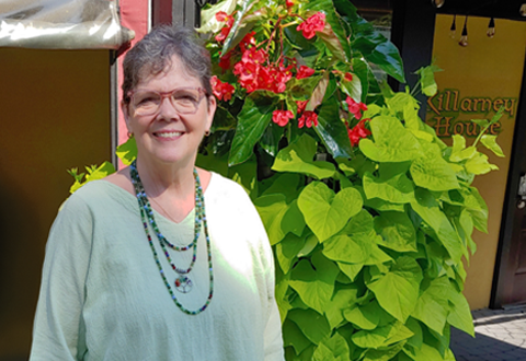 A woman with short hair and glasses, wearing a light green top and beaded necklaces, stands smiling in front of a lush green plant with red flowers outside a building with a sign that reads “Killarney House”