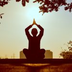 Silhouette of a person sitting with crossed legs and hands pressed together over head. 