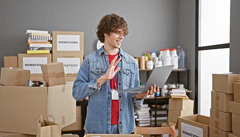 A friendly young man with curly hair stands amid cardboard boxes in a warehouse, using a laptop and gesturing.